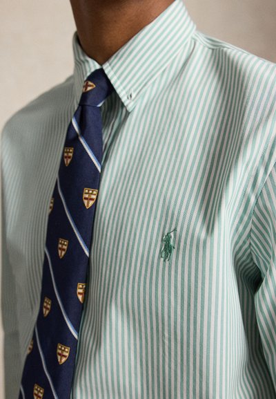 Homme portant une chemise à rayures vertes et blanches boutonnée avec une cravate bleu marine avec des emblèmes de bouclier dorés.