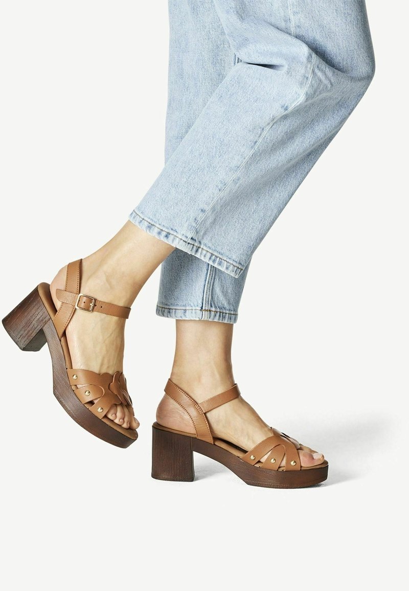 Des pieds portant des sandales à talons en cuir beige avec des brides de cheville, associées à un jean en denim bleu clair coupé.