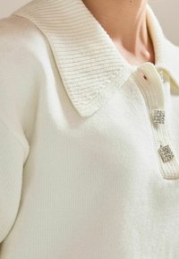 Cremefarbener Pullover mit einem gerippten Kragen, drei dekorativen Knopfverschlüssen und einem strukturierten Stoff mit weichem Finish.