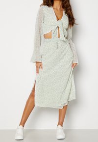 Vestido floral verde claro con mangas largas, un lazo en el frente y una abertura lateral, hecho de una tela transparente. Combinado con zapatillas blancas.