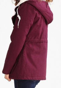 Bordeauxfarvet hættetrøje med hvid fleeceforing, justerbar snøre og en afslappet pasform. Har en mellemlang længde og sidelommer.