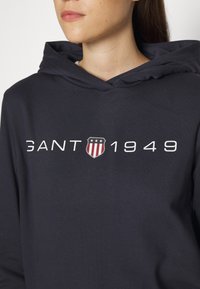Femme portant un sweat à capuche foncé avec un logo en forme de bouclier et le texte "GANT 1949" sur la poitrine.