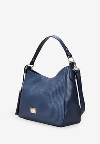 Bolso de mano azul marino con un patrón texturizado. Cuero suave, acentos de hardware en dorado y una correa desmontable. Forma curva con un detalle de borlas.