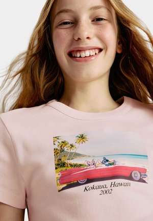Roze katoenen T-shirt met een afbeelding van twee cartoonfiguren in een rode cabriolet, omringd door palmbomen en een strandachtergrond, met de tekst "Kokaua, Hawaii 2002."