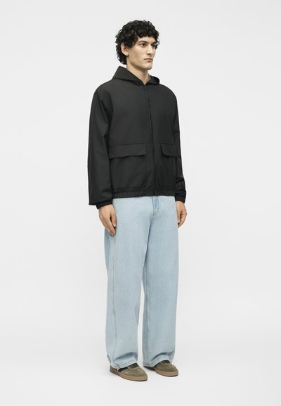 Νεαρός άνδρας με σγουρά μαλλιά φοράει μαύρο μπουφάν με κουκούλα, ανοιχτό γαλάζια φαρδιά τζιν και καφέ αθλητικά παπούτσια, στέκεται μπροστά σε λευκό φόντο.