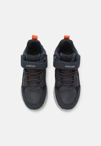 Zapatillas altas azul marino con acentos naranjas, que presentan una correa de Velcro y cordones. Superficies suaves y texturizadas, suelas de goma.