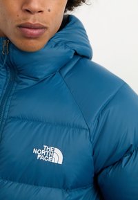 Mėlynas pūkinis džemperis su gobtuvu, turintis siuvinėtą dizainą, užtrauktuką ir baltą "THE NORTH FACE" logotipą ant krūtinės. Sklandus paviršius.