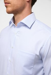 Camicia azzurra chiara con bottoni, realizzata in tessuto testurizzato, con colletto a punta, taschino sul petto e bottoni bianchi.