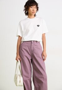 Biała bawełniana koszulka z czarnym logo w kształcie serca, zestawiona z fioletowymi spodniami z wysokim stanem i małą białą torebką. Jednolite tło.