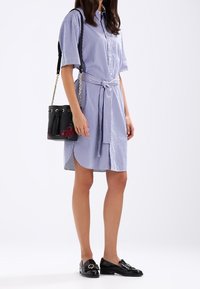 Randig skjortklänning i ljusblått och vitt, åtdragen i midjan, med en svart buckettväska som har blommiga detaljer och en tofs.