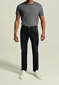 Mężczyzna w szarej koszulce z krótkim rękawem, czarnych obcisłych dżinsach i białych tenisówkach, stojący na jednolitym tle.