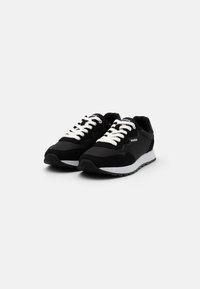Baskets de sport noires avec un dessus texturé, semelle blanche contrastante et lacets en nylon. Présente un logo sur la languette et sur le côté.