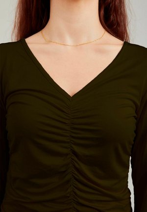 Sieviete, kas valkā tumši zaļu rušētu V veida izgriezuma garām piedurknēm topu un plānu zelta ķēdītes kaklarotu, redzama no kakla līdz viduklim.