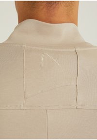 Beige Stoffoberteil mit hohem Kragen, sichtbaren Nähten und einer gestickten Detail in einem helleren Beige im Bereich des Nackens.