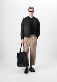 Zwarte bomberjack over een zwart t-shirt, beige wijde broek, zwarte leren handtas, zwarte sandalen en een zonnebril.