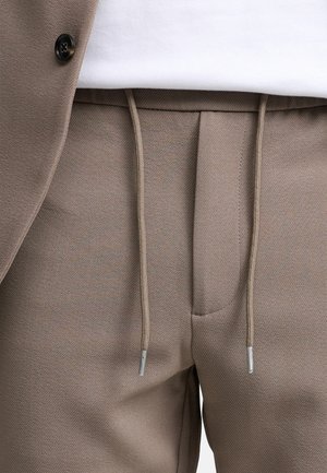 Beżowe dresy z teksturowanej tkaniny, elastycznym pasem i detalami ze sznurkiem. Wykończenie obejmuje srebrne elementy metalowe i subtelny wzór w jodełkę.