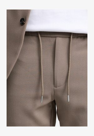 Beżowe dresy z teksturowanej tkaniny, elastycznym pasem i detalami ze sznurkiem. Wykończenie obejmuje srebrne elementy metalowe i subtelny wzór w jodełkę.