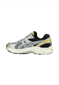 Zapatilla de correr plateada con parte superior de malla negra, acentos amarillos y suela blanca acolchada. Presenta un diseño en capas y texturas contrastantes.