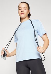 Camiseta deportiva de manga corta de color azul claro con un branding sutil, combinada con leggings negros. El modelo sostiene una cuerda de saltar con mangos grises.