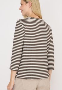 Femme aux longs cheveux blonds portant un haut à rayures horizontales marron et blanc à manches trois-quarts et un pantalon beige, orientée vers la gauche.