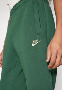 Zelené tepláky vyrobené z měkkého materiálu s elastickým pasem, boční kapsou a zlatým logem Nike na stehně.
