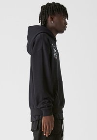 Fekete pamut kapucnis pulóver kenguruzsebbel, bordázott mandzsettákkal és grafikai nyomattal elöl. Puha textúra és laza szabás. 