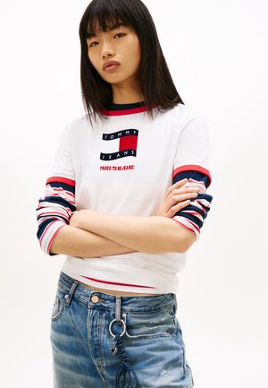 Ung kvinde med glat sort hår iført hvid Tommy Jeans skjorte med røde og blå stribede ærmer og slidte blå jeans med nøglering.