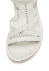 Sandal in pelle bianca con cinturini incrociati. Il design presenta cuciture visibili e una suola piatta, con un messaggio stampato all'interno.