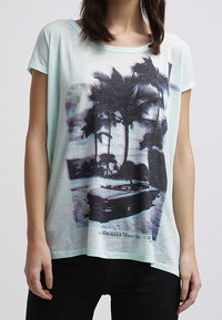 Mintgrøn t-shirt lavet af blødt stof med et grafisk print af palmer og en bil. Afslappet pasform med rund halsudskæring og korte ærmer.