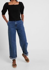 Černý top s nabíranými rukávy a čtvercovým výstřihem, doplněný o volné modré džíny s ohrnutými manžetami a hnědé espadrilové boty na klínu.