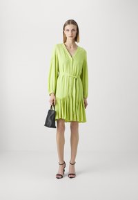 Lime green dress with a V-neck, long puff sleeves, button front, and a belted waist. The skirt has a ruffled hem. Paired with a black handbag.