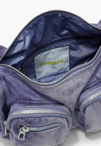 Ljubičasta platnena torba s sjajnom unutrašnjošću, s dva zipovana džepa, oznakom branda u žutoj boji i izvezenim logotipom.