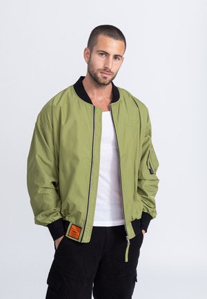 Olijfgroene bomberjack met een zwarte kraag, ritssluiting, geribde manchetten en een patch op de linkermouw. Gemaakt van lichtgewicht materiaal.