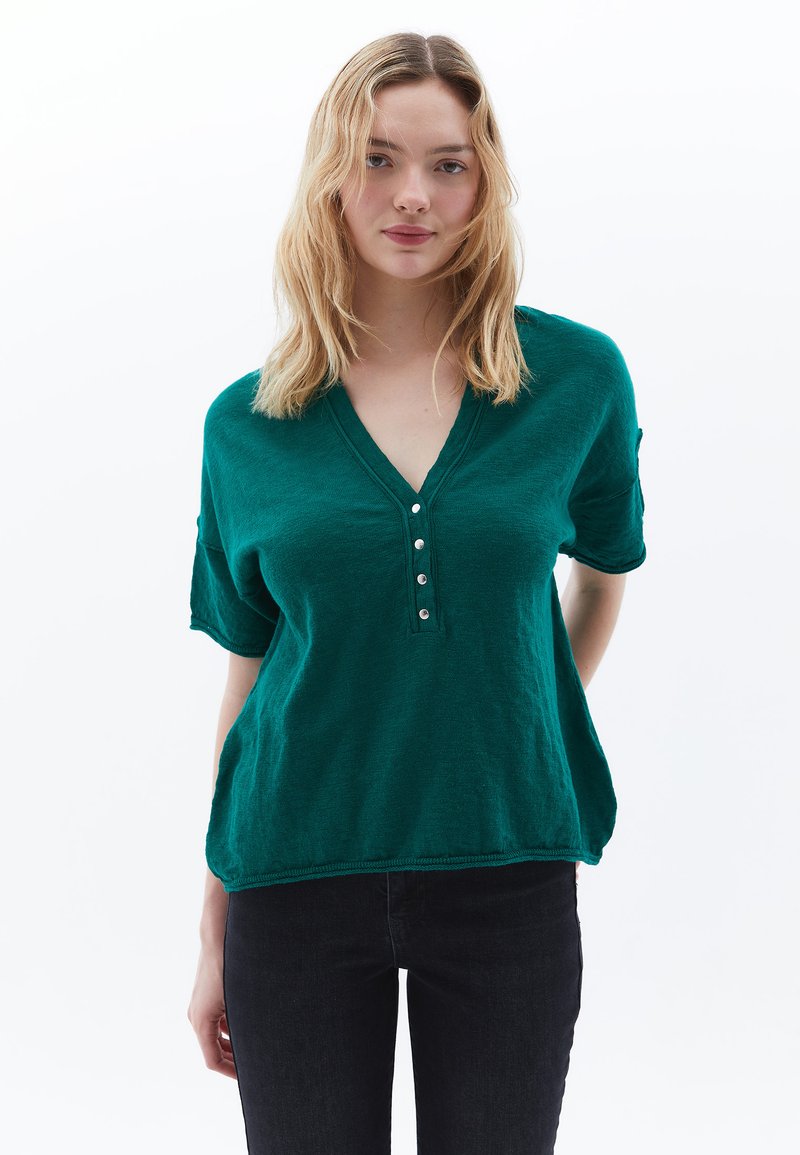 Teal katoenmengsel korte mouwen top met een v-hals, met knoopsluiting en omgerolde mouwen, gecombineerd met donkere jeans. Zachte textuur.