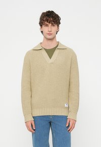 Beżowy dziergany sweter z dekoltem w serek i szerokim kołnierzem. Ściągacze na mankietach i u dołu. Nałożony na oliwkowozieloną koszulkę. Lekka faktura.