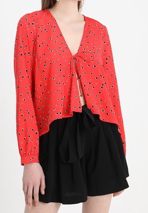 Blusa rossa con motivi circolari neri e bianchi, scollatura a V, lacci davanti e maniche lunghe. Abbinata a pantaloni corti neri.