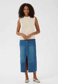 Top sans manches beige associé à une jupe midi en denim bleu avec une fente avant. La tenue est complétée par des chaussures plates de couleur claire.