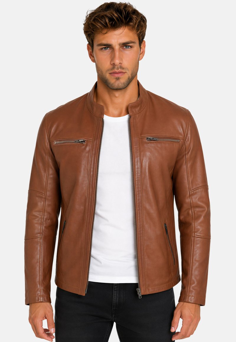 Jonge man met kort krullend haar, gekleed in een bruine leren jas over een wit overhemd en een zwarte broek, staand tegen een effen achtergrond.