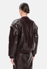 Giubbotto bomber in pelle marrone con orlo arricciato, maniche oversize e dettagli con cuciture verticali. Texture morbida e lucida.