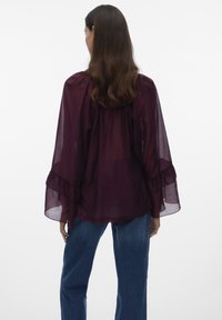 Frau mit langem braunem Haar trägt eine transparente violette Bluse mit gekräuselten Glockenärmeln und blaue Jeans, von hinten vor weißem Hintergrund gezeigt.