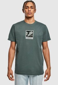 T-shirt di cotone verde scuro con colletto rotondo, maniche corte e una grafica frontale che mostra la parola "glamorous" all'interno di un bordo quadrato.