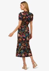 Vrouw in een kleurrijke bloemen midi-jurk met puffmouwen en een gerimpelde zoom, die gouden hoge hakken draagt, in profiel staand tegen een witte achtergrond.