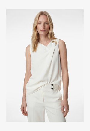 Blonde woman wearing a sleeveless white top with a gold circular clasp holding a draped fold near the shoulder, paired with white pants.