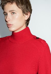 Osoba s krátkými vlasy, která má na sobě texturovaný červený rolák s knoflíkovými ramenními popruhy, dívající se vlevo na jednotném pozadí.