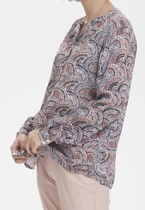 Personne portant un chemisier à manches longues avec des motifs circulaires multicolores et un pantalon rose clair, ajustant l’ourlet du chemisier avec les mains.