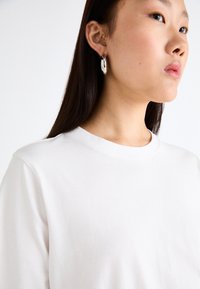 T-shirt en coton blanc avec un col rond, accompagné de boucles d'oreilles argentées en forme de créole. Texture lisse avec un léger éclat sur les bijoux.