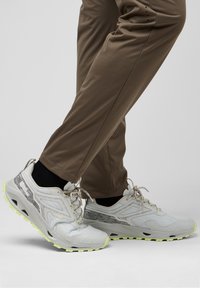 Lichtgrijze sportschoenen met een gestructureerde bovenkant, reflecterende accenten en een neonkleurige gele loopzool; gecombineerd met strakke bruine broeken.