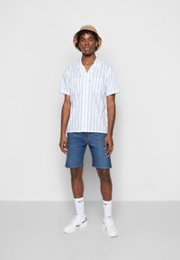 Camisa de manga curta às riscas em azul claro e branco, combinada com calções de ganga. O modelo usa um chapéu de aba larga bege e ténis brancos.