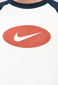 Κοντινό πλάνο ενός λευκού και ναυτικού μπλουζιού raglan με ένα κόκκινο οβάλ που περιέχει το λευκό λογότυπο Nike swoosh στο στήθος.