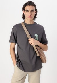 Camiseta gris de manga corta con puños doblados, que presenta un diseño gráfico verde en el frente. La persona sostiene un bolso de hombro marrón claro.